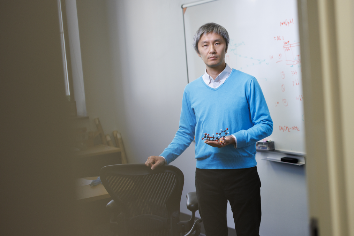 Ken Yokoyama stands in front of a whiteboard holding a model of a molecule.